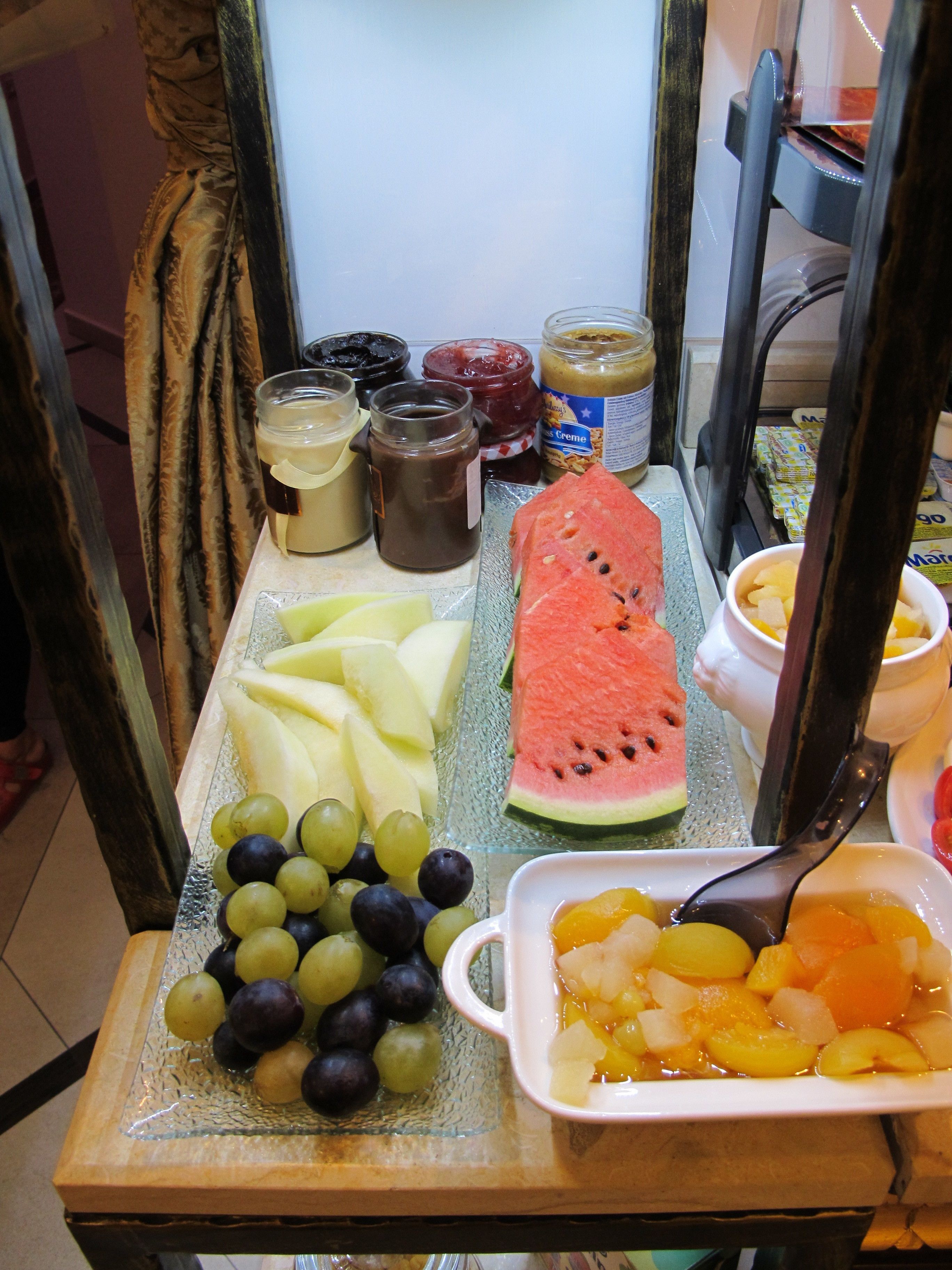 A table with a variety of fruits, including watermelon, apples, grapes, and oranges, along with several jars of spreads and condiments.