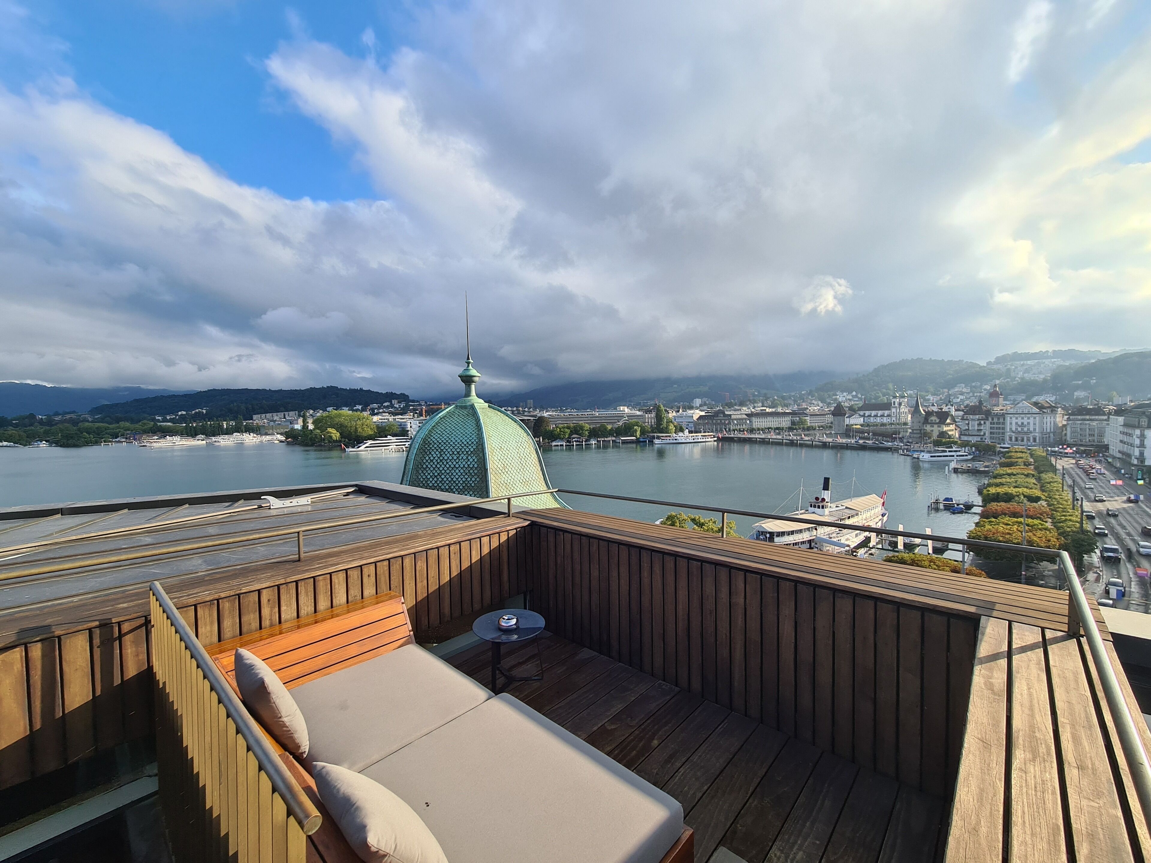 A wooden deck with a sofa and table, overlooking a river with a cityscape and a distinctive green-domed building in the background.