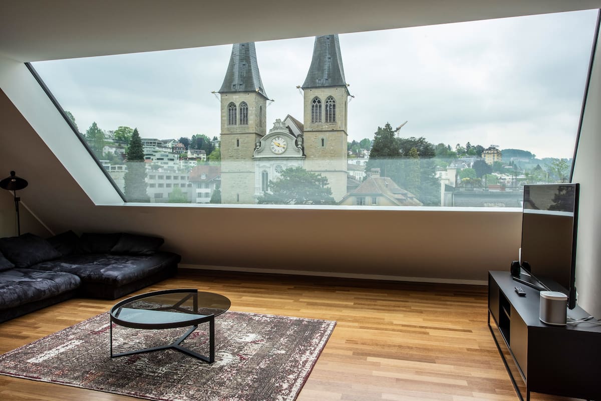 A modern loft bedroom with a skylight, a black sofa, a glass coffee table, and a view of a church tower.
