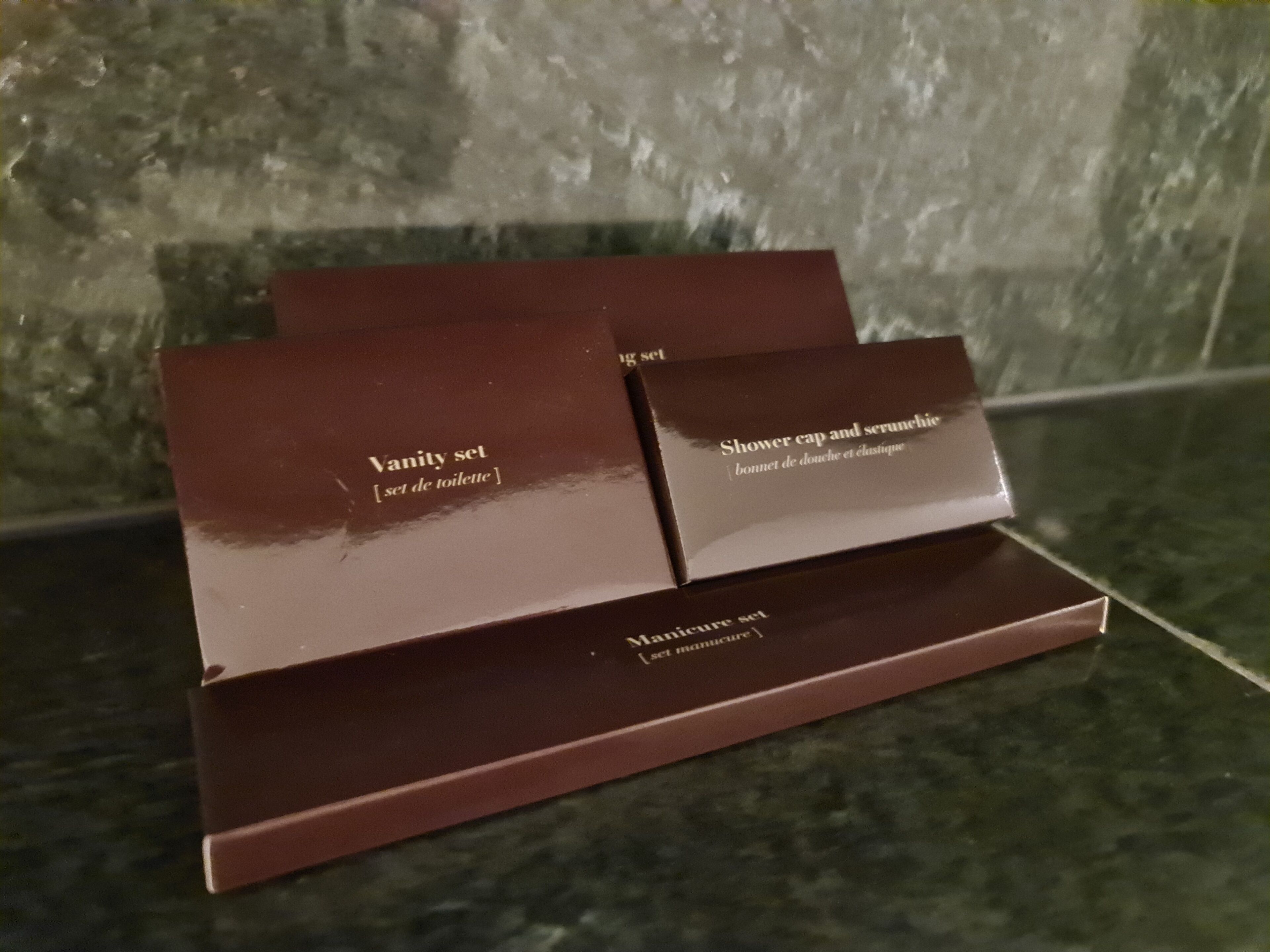 Tres cajas marrones etiquetadas 'Vanity set', 'Gorro de ducha y seruniche' y 'Set de manicura' sobre una superficie de mármol.