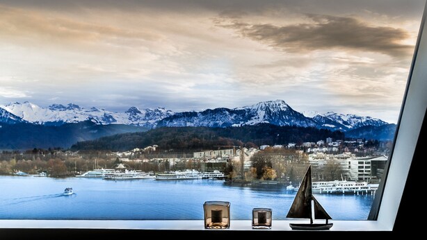 A view from a window overlooking a lake with mountains in the background, a cruise ship, and a small sailboat model on the windowsill. - Lucerne Lake View Apartments (Lucerne)