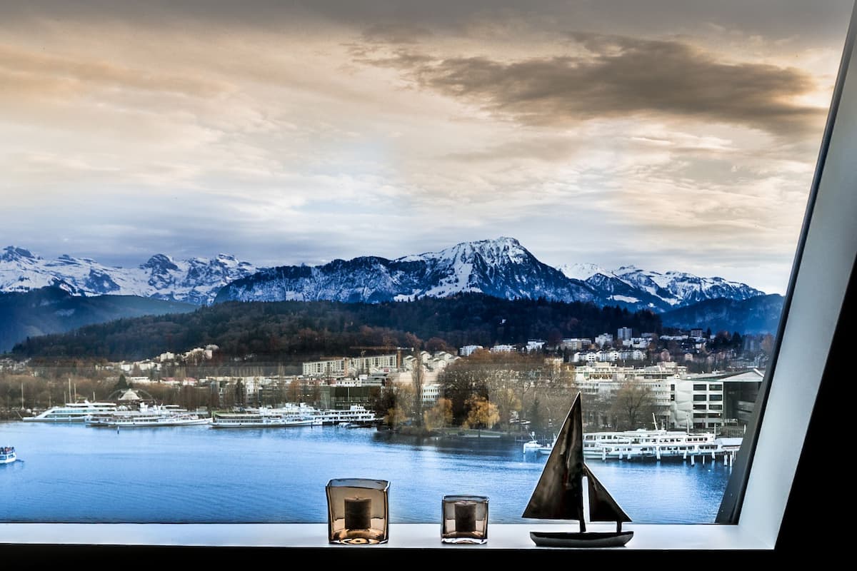 A view from a window overlooking a lake with mountains in the background, a cruise ship, and a small sailboat model on the windowsill.