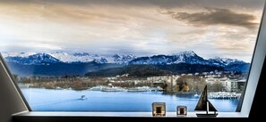 A view from a window overlooking a lake with mountains in the background, a cruise ship, and a small sailboat model on the windowsill. - Lucerne Lake View Apartments (Lucerne)