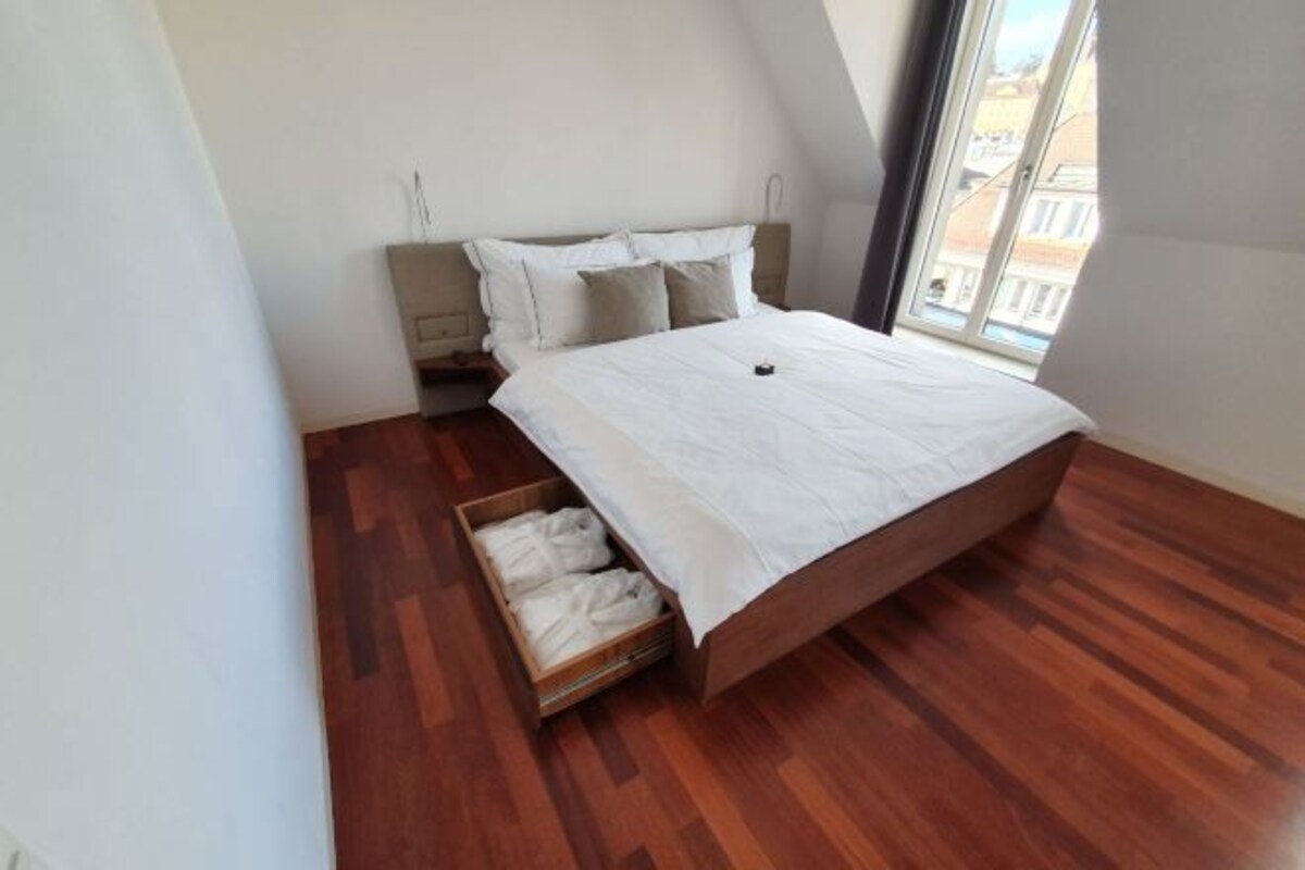 A bedroom with a wooden floor, a bed with white bedding, a wooden nightstand, and a window with a view of buildings.