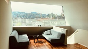A modern room with a large window offering a view of a cityscape, including buildings and a hill. There are two grey sofas and a small round table in the center. - Lucerne Lake View Apartments (Lucerne)