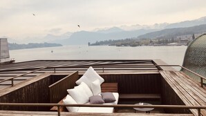 A wooden deck with a cushioned seating area overlooking a lake with boats and mountains in the background. - Lucerne Lake View Apartments (Lucerne)