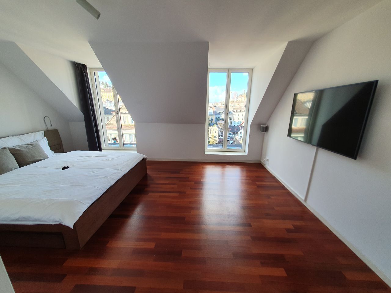Ein Schlafzimmer mit einem Bett, Holzboden, einem Fernseher und einem großen Fenster mit Blick auf die Stadt.