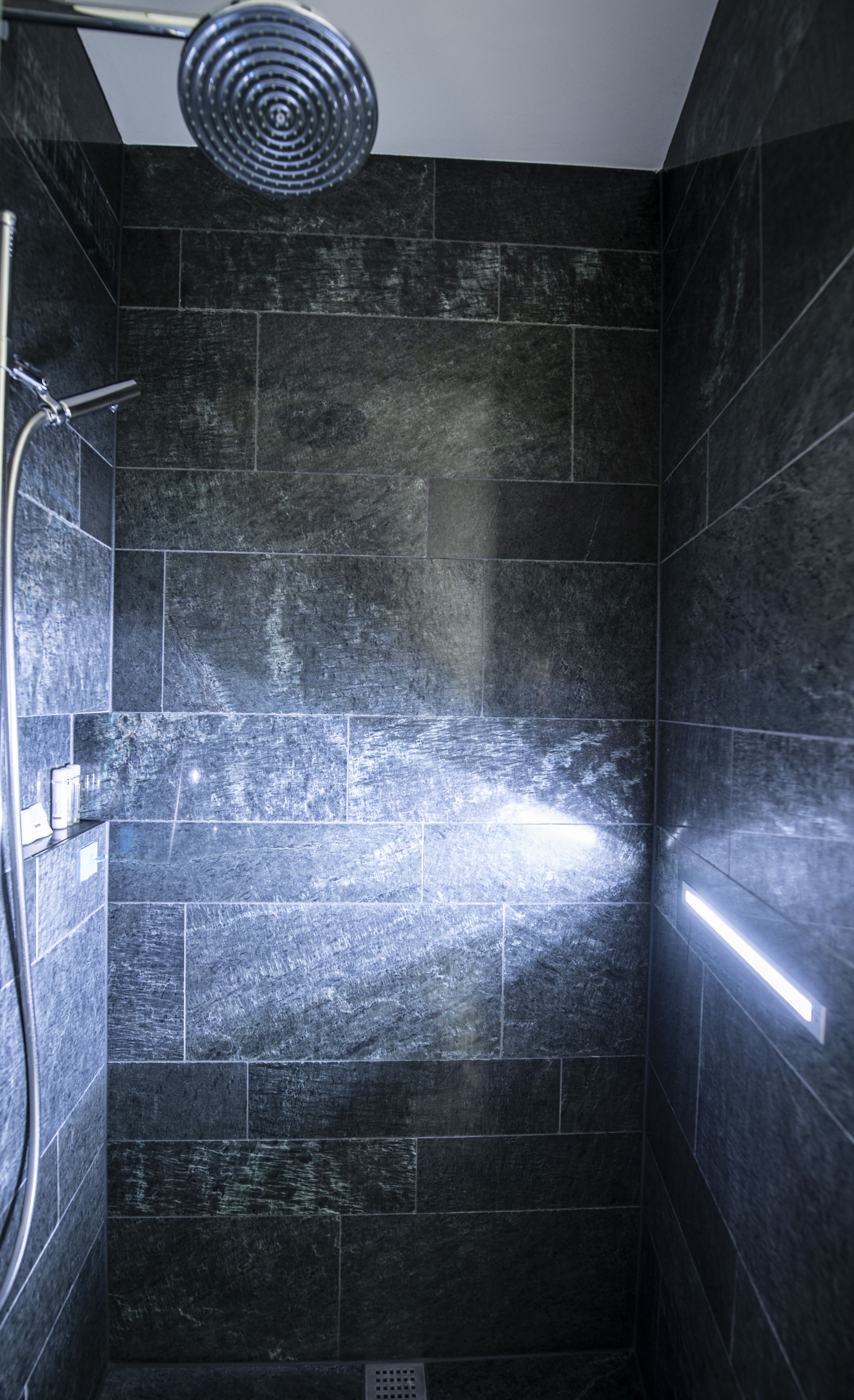 Ducha con un elemento acuático circular, paredes de azulejos oscuros y desagüe visible.