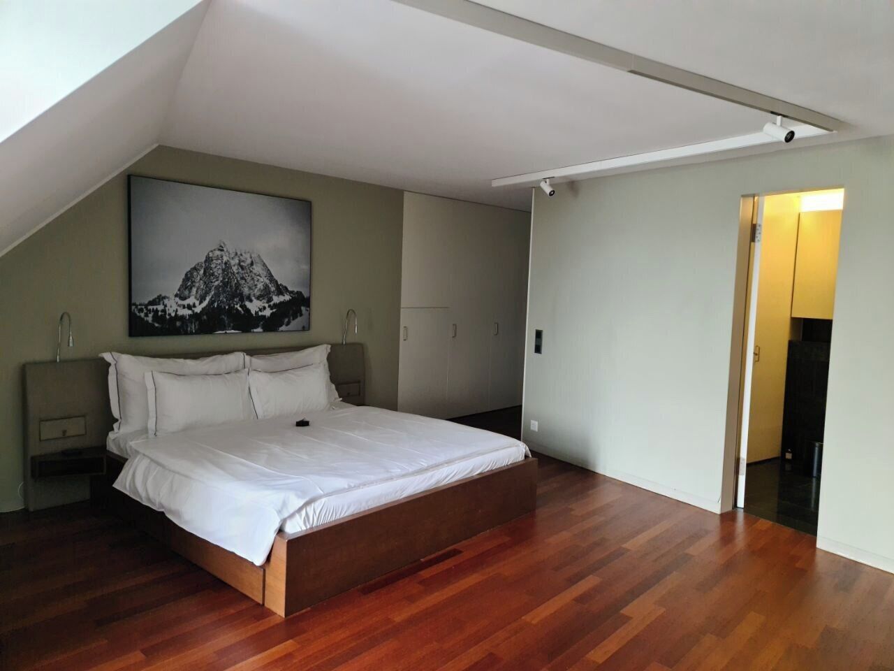 Un dormitorio con piso de madera, una cama con ropa blanca, una mesita de noche, un estante montado en la pared y un cuadro de una montaña.