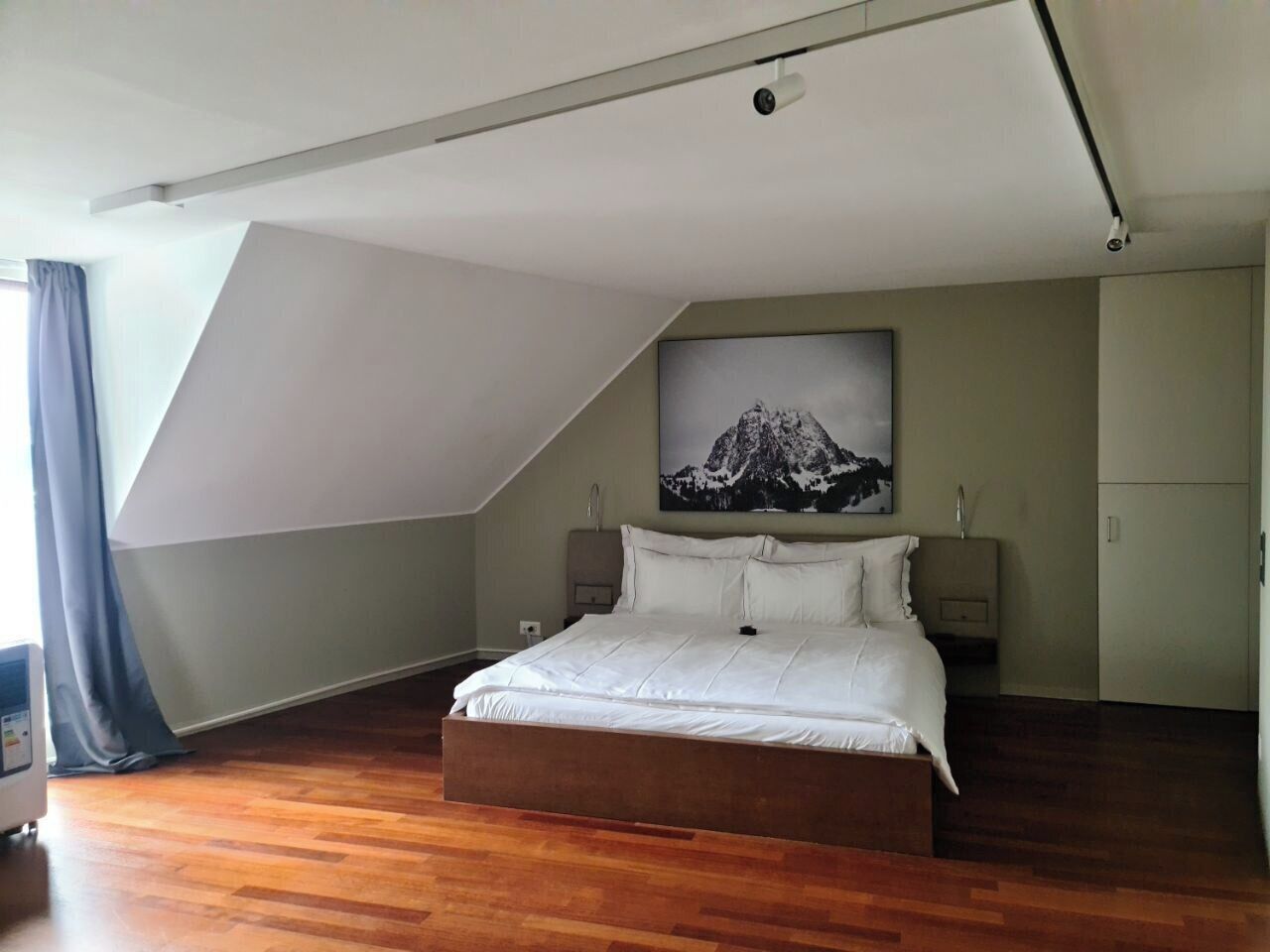 Un dormitorio con techo inclinado, una cama, una mesita de noche, un televisor montado en la pared y un cuadro de una montaña.