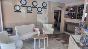 Un hall de hotel con un sofá, sillas, una mesita, relojes mostrando diferentes horas de ciudades y una recepción, además de una chimenea.