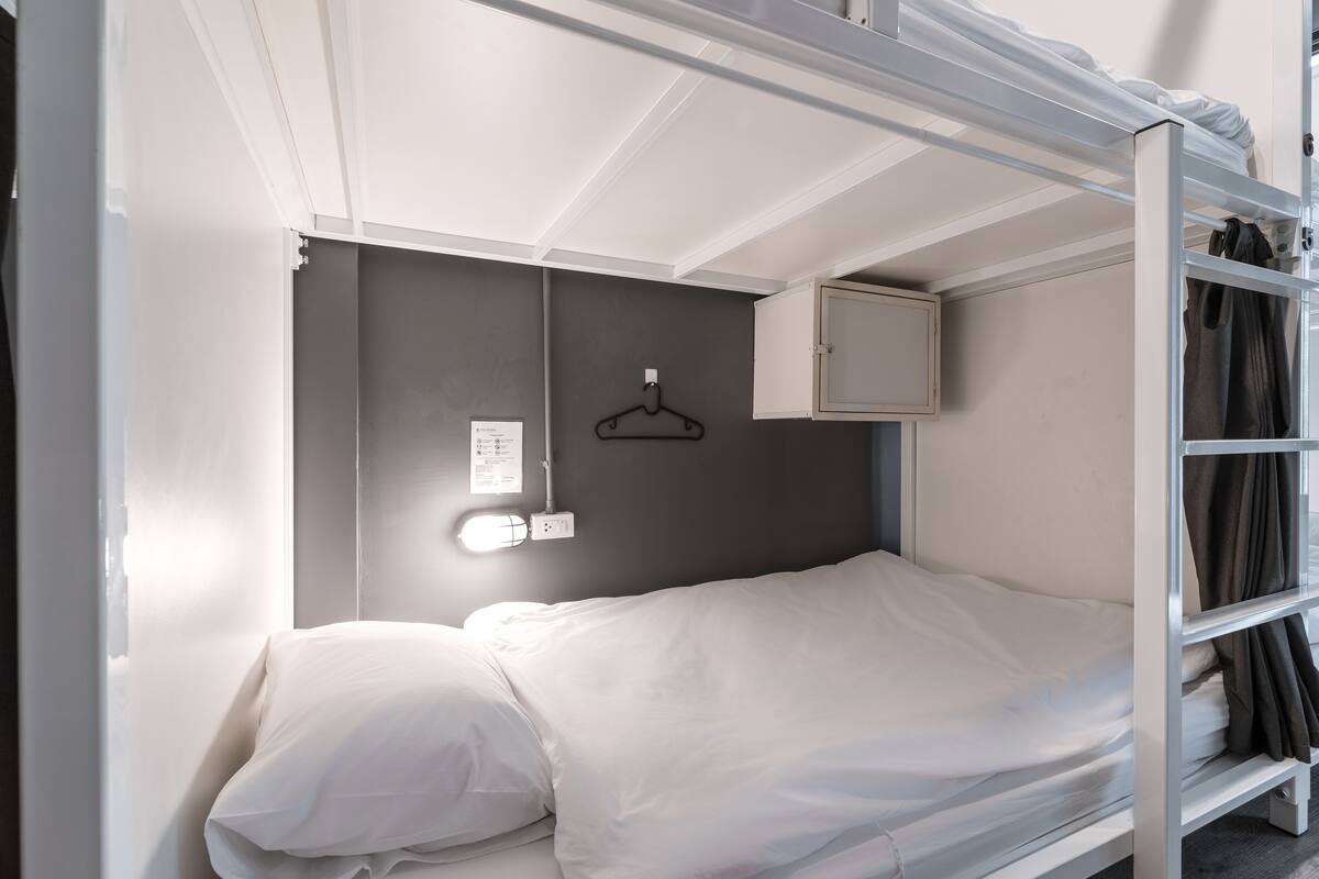 A bunk bed with a white mattress and pillows, a small cabinet, and a wall-mounted light fixture.