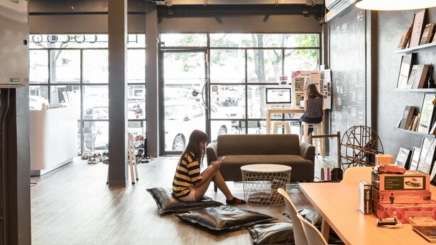 A modern interior with a sofa, a round table, and a person sitting on a cushion. - The Cocoon Hostel (Bangkok)
