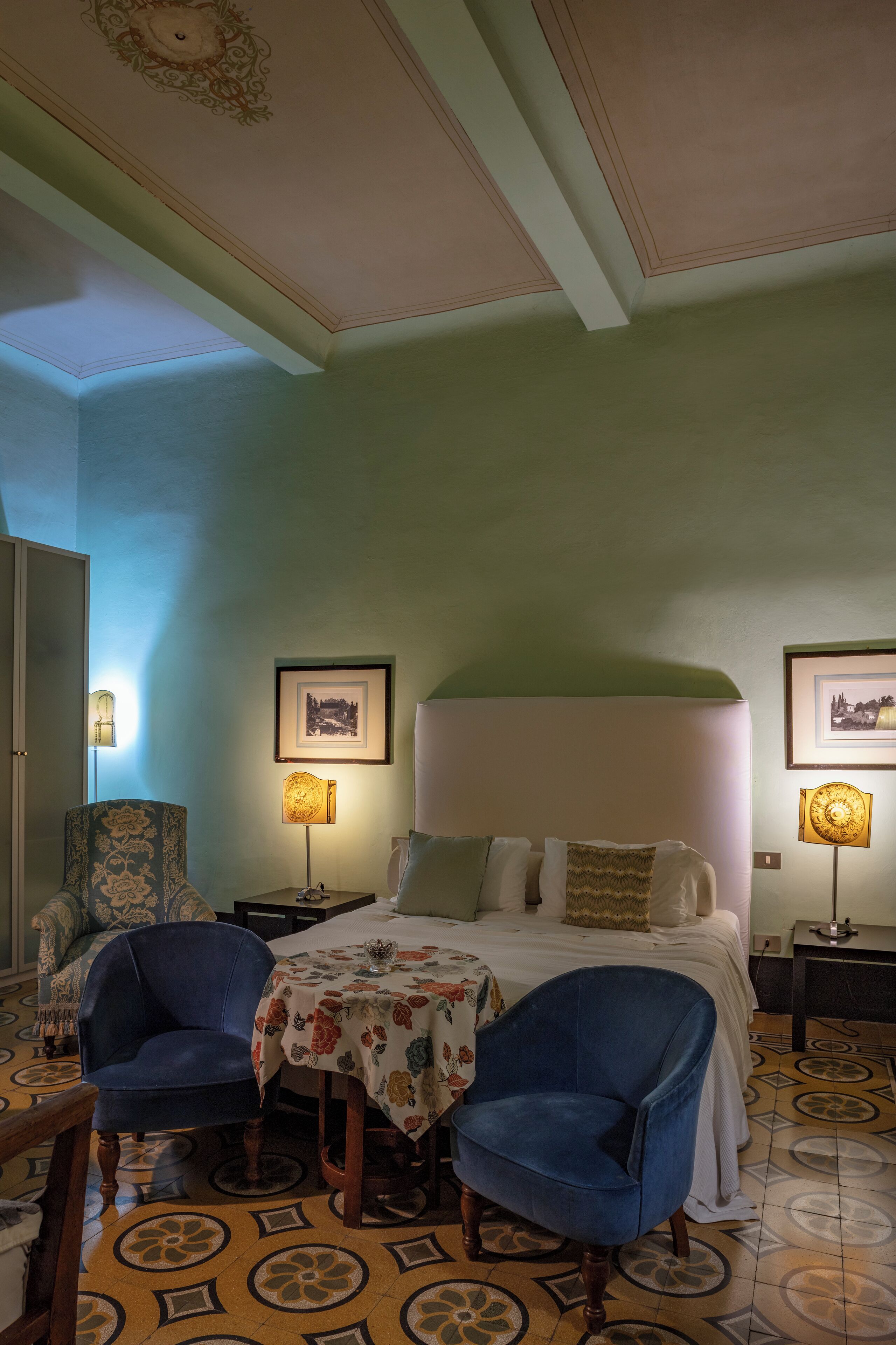 A hotel room with a bed, two chairs, a table, and framed pictures on the walls.