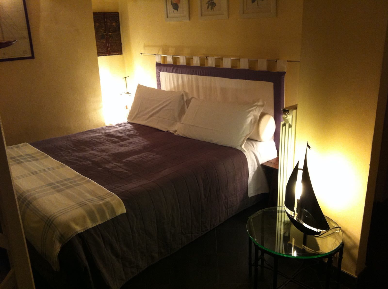 A neatly made bed with a purple and white duvet, a glass side table with a lamp, and a framed picture on the wall.