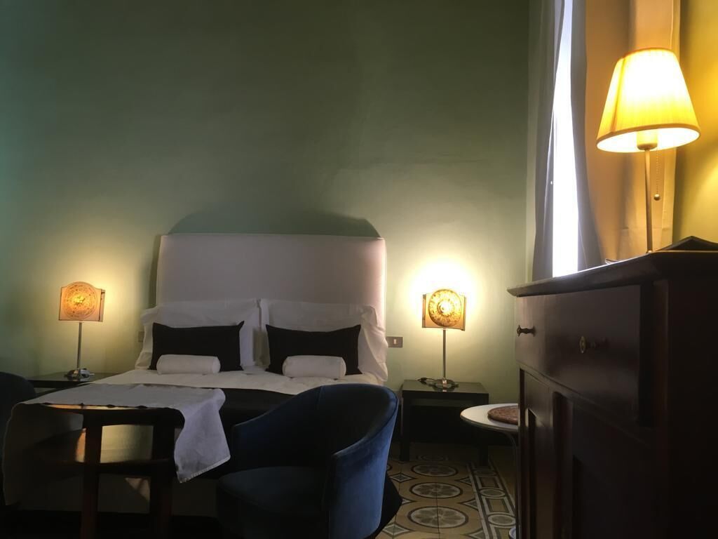 Una habitación de hotel con una cama, dos lámparas de noche, una silla, un tocador y una mesita.