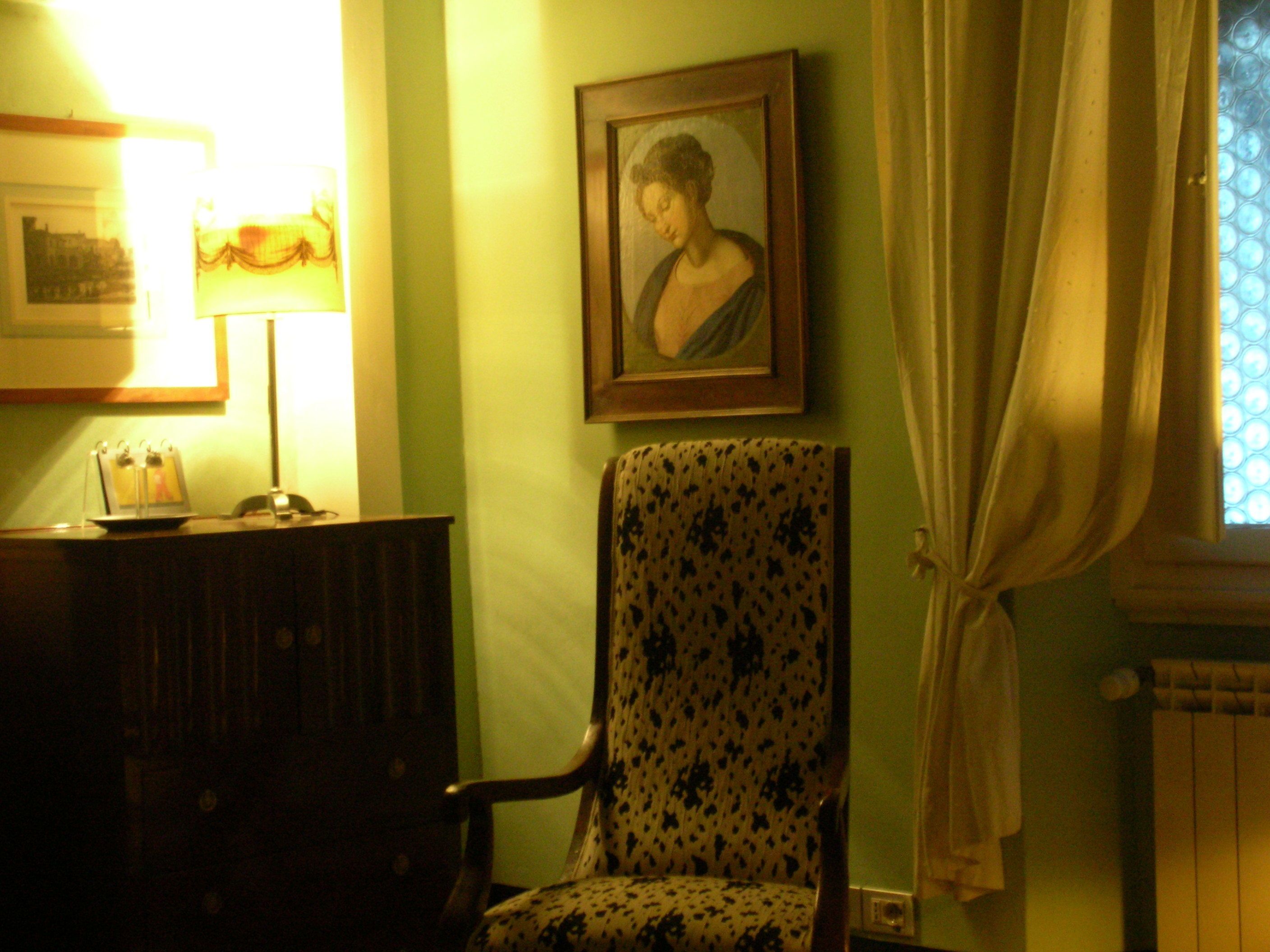 A room with a patterned armchair, a framed portrait on the wall, a desk with a lamp, and a window with curtains.