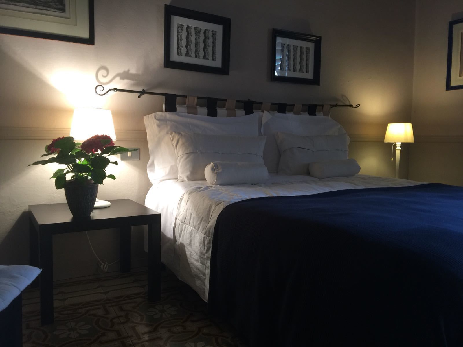 A neatly made bed with white bedding and a dark blue blanket, flanked by two bedside lamps. A small table with a potted plant is positioned to the left of the bed.