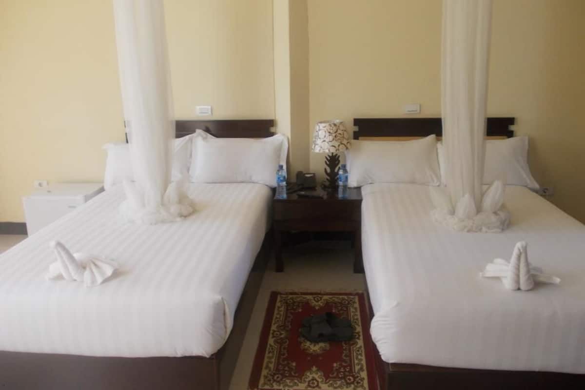 A hotel room with two beds, each with white linens and a canopy. There is a nightstand between the beds with a lamp and a vase of flowers.