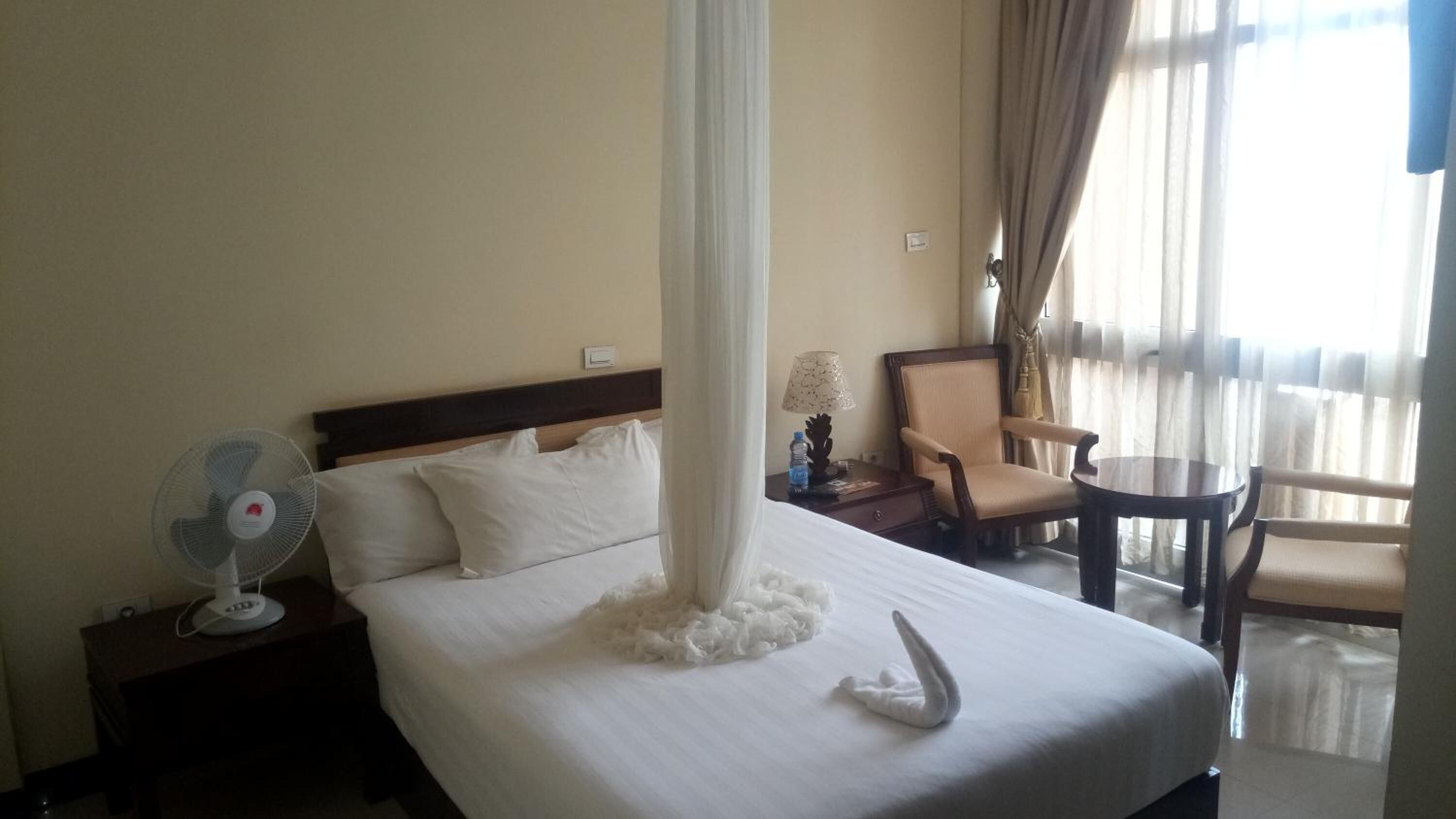 A hotel room with a bed, a bedside fan, a chair, a table, and a window with curtains.