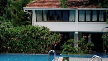 A swimming pool with a tiled deck, surrounded by lush greenery and a building with a tiled roof.