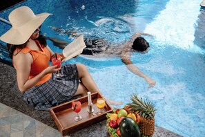 Una persona leyendo un libro junto a una piscina, con una bandeja de bebidas y frutas.