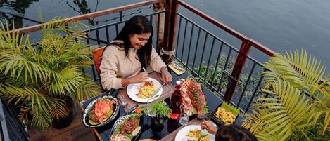 A dining area with a view of water, featuring a table with various dishes and a bottle of red beverage.