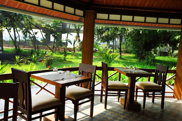 An outdoor dining area with wooden tables and chairs, surrounded by lush greenery and a view of a garden.