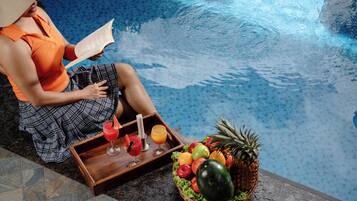 Una persona sentada junto a una piscina, con una bandeja de bebidas y una cesta de frutas.