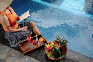 Una persona sentada junto a una piscina, con una bandeja de bebidas y una cesta de frutas.