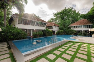 A swimming pool with a tiled deck, surrounded by lush greenery and two buildings with tiled roofs.