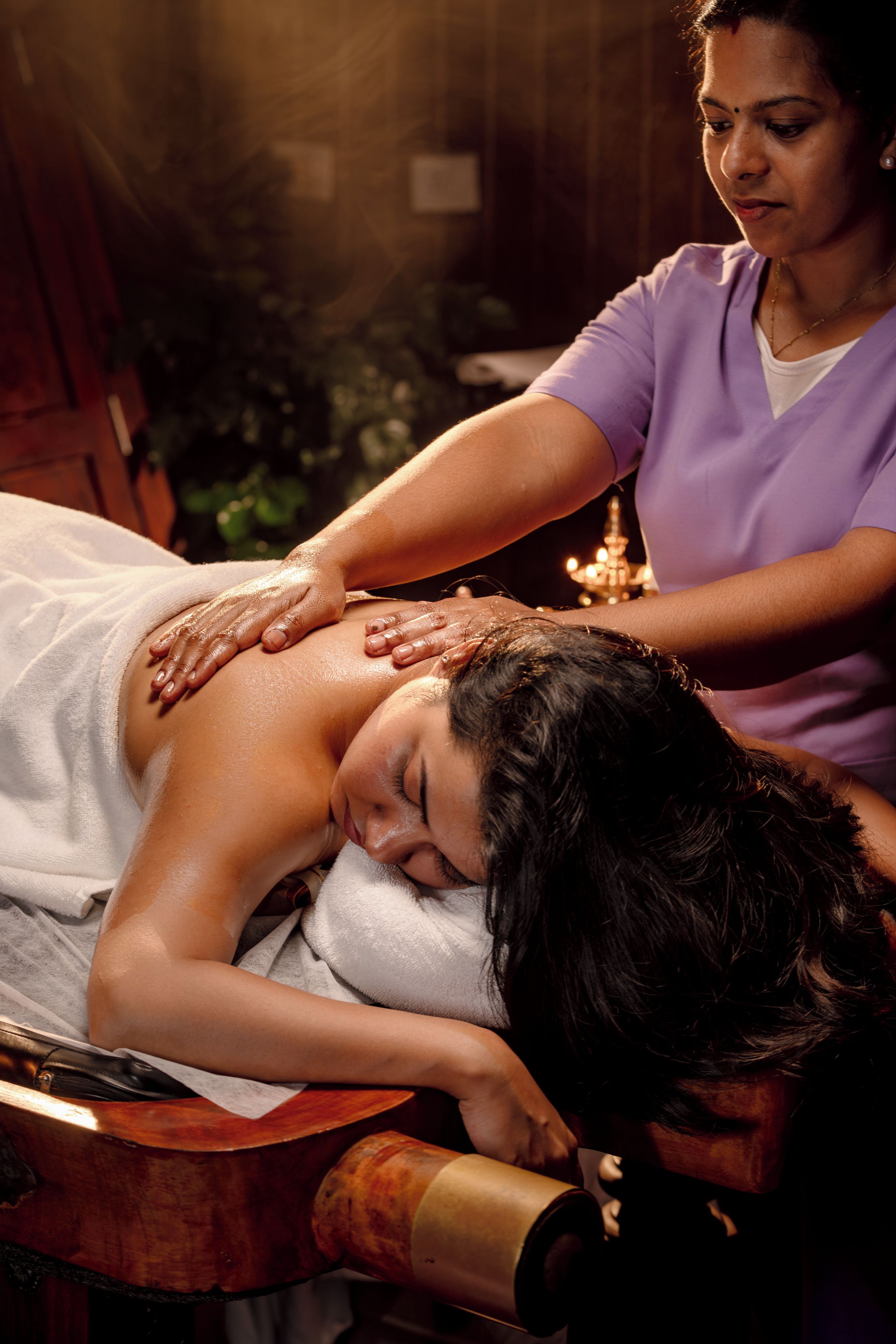 A massage therapy session in a room with a wooden massage table, a candle, and a plant in the background.