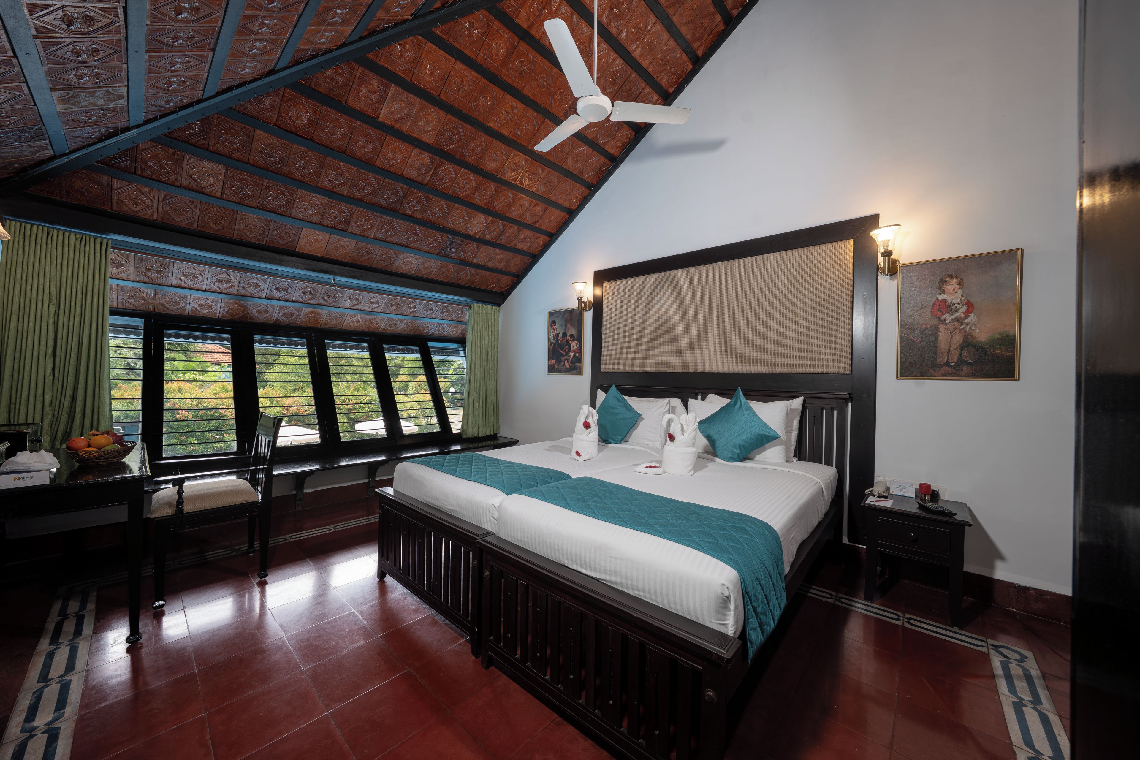 Habitación de hotel con cama, mesita de noche, ventana con cortinas, ventilador de techo y un cuadro en la pared.
