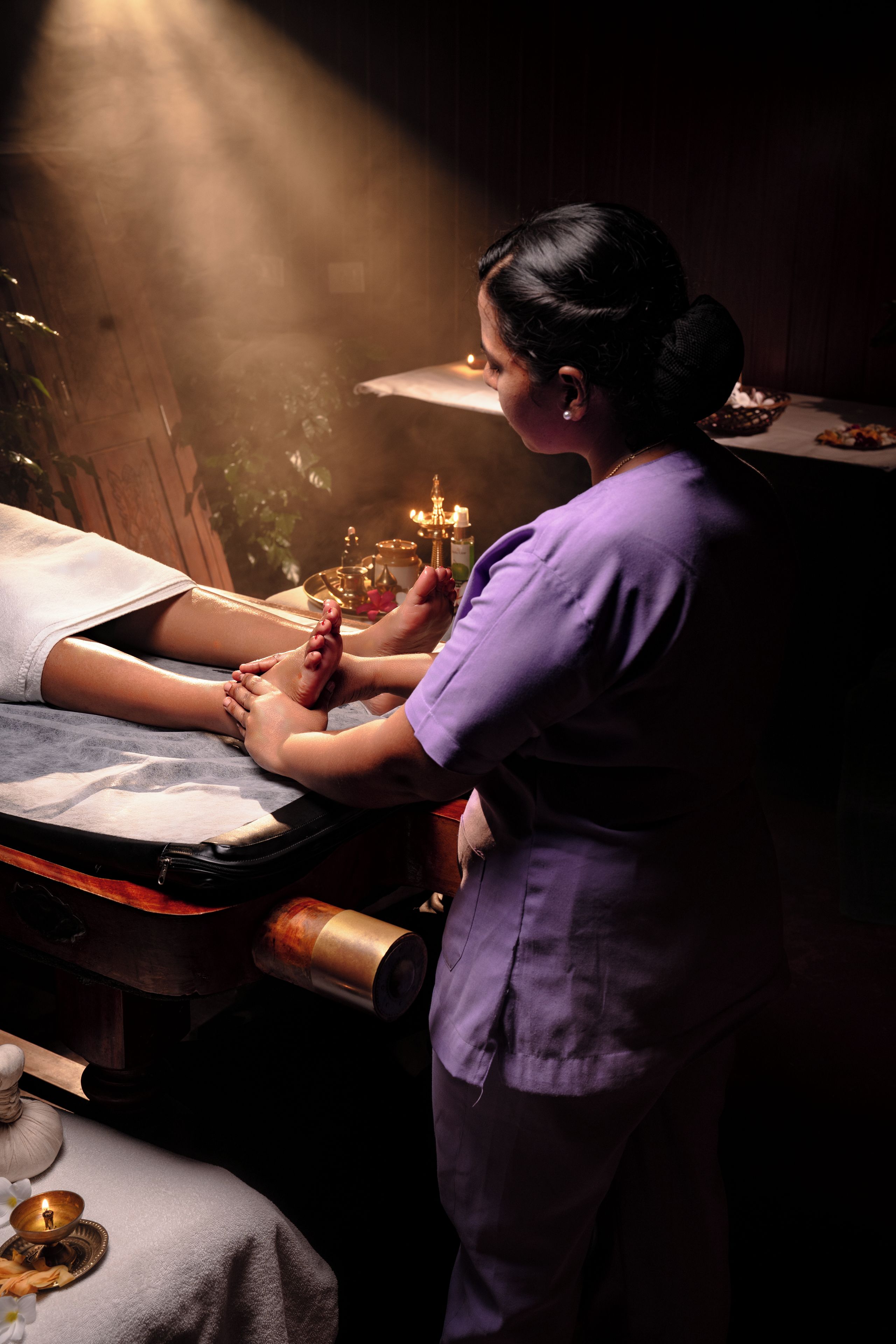 A person receiving a foot massage in a dimly lit room with a massage table, a candle, and a small table with items.