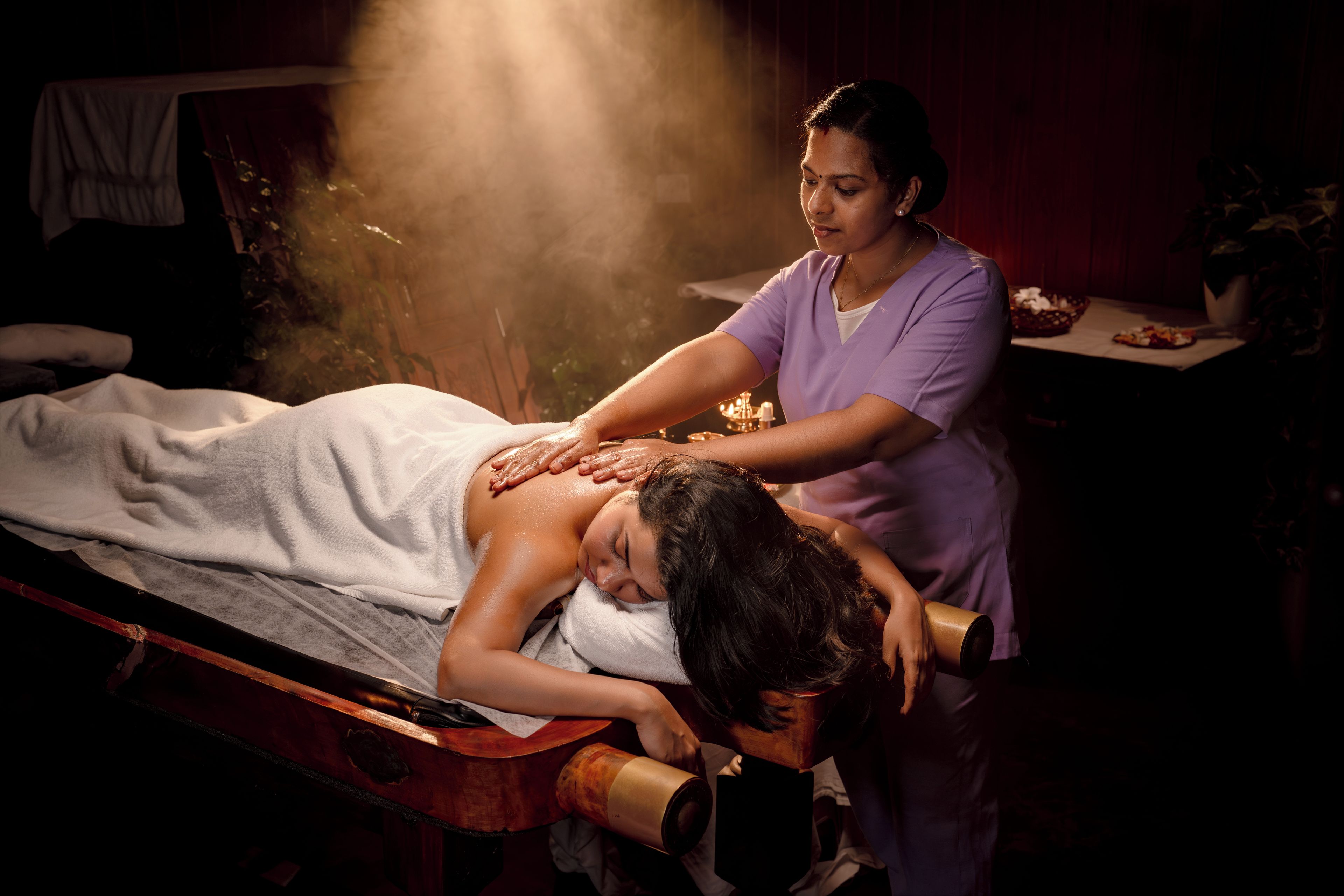 A massage therapy session in a dimly lit room with a massage table, a person receiving a back massage, and a steam treatment device.