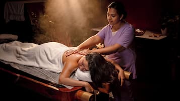 Sessão de massagem em ambiente com pouca luz, com maca de massagem, uma pessoa recebendo massagem nas costas e um aparelho de vaporização.