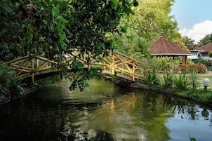 A bamboo bridge over a calm water body, surrounded by lush greenery and a traditional building with a tiled roof.