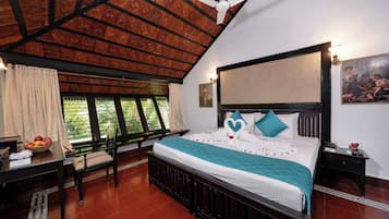 Habitación de hotel con cama, escritorio, silla, ventana con cortinas, ventilador de techo y un cuadro en la pared.