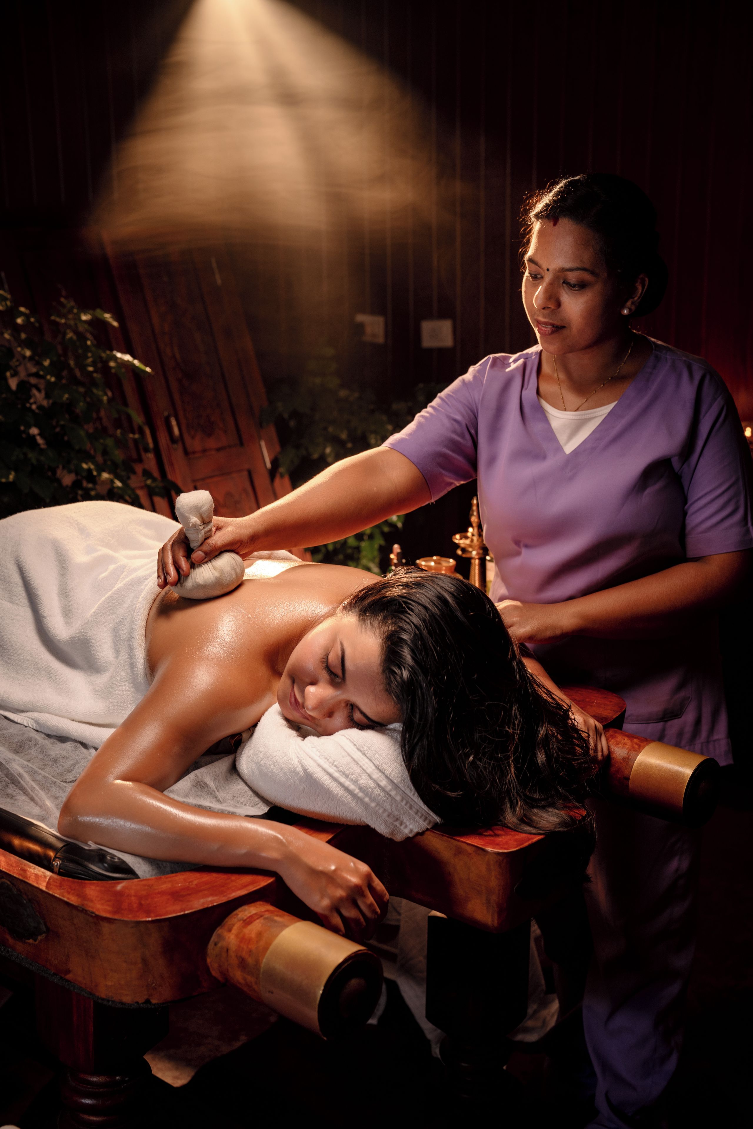 A person receiving a massage in a dimly lit room with a wooden massage table and a massage chair.
