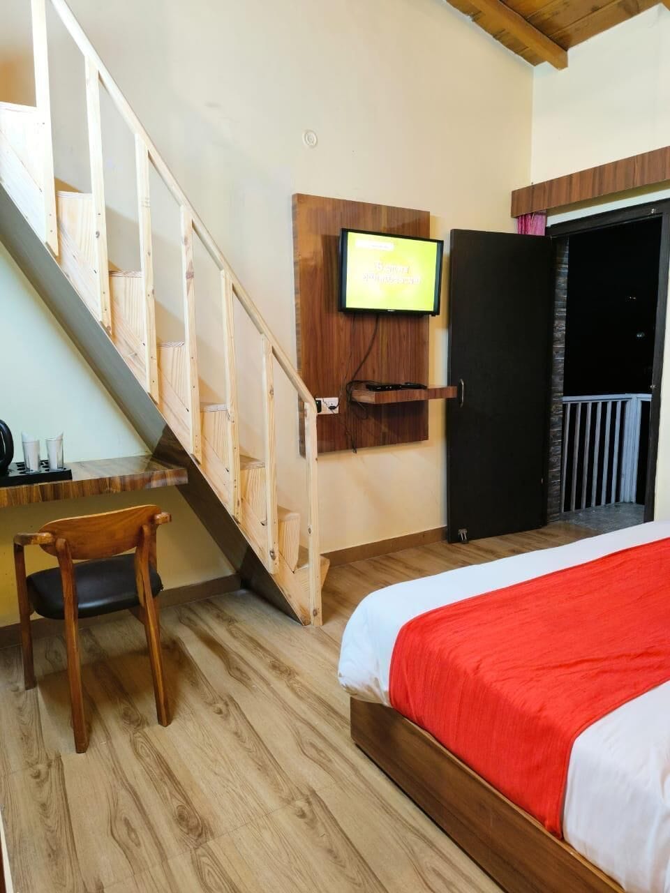 Ein Schlafzimmer mit einer Holztreppe, einem Bett mit roter Decke, einem an der Wand montierten Fernseher und einer Tür, die auf einen Balkon führt.
