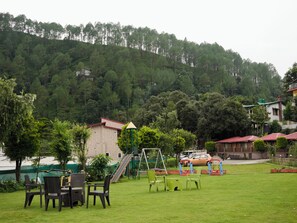 A grassy outdoor area with a playground, including swings and slides, surrounded by trees and buildings.