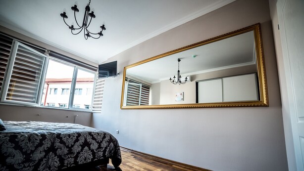 A bedroom with a large mirror, a bed with a patterned bedspread, wooden flooring, and a chandelier. - Suite Donna (Bihac)