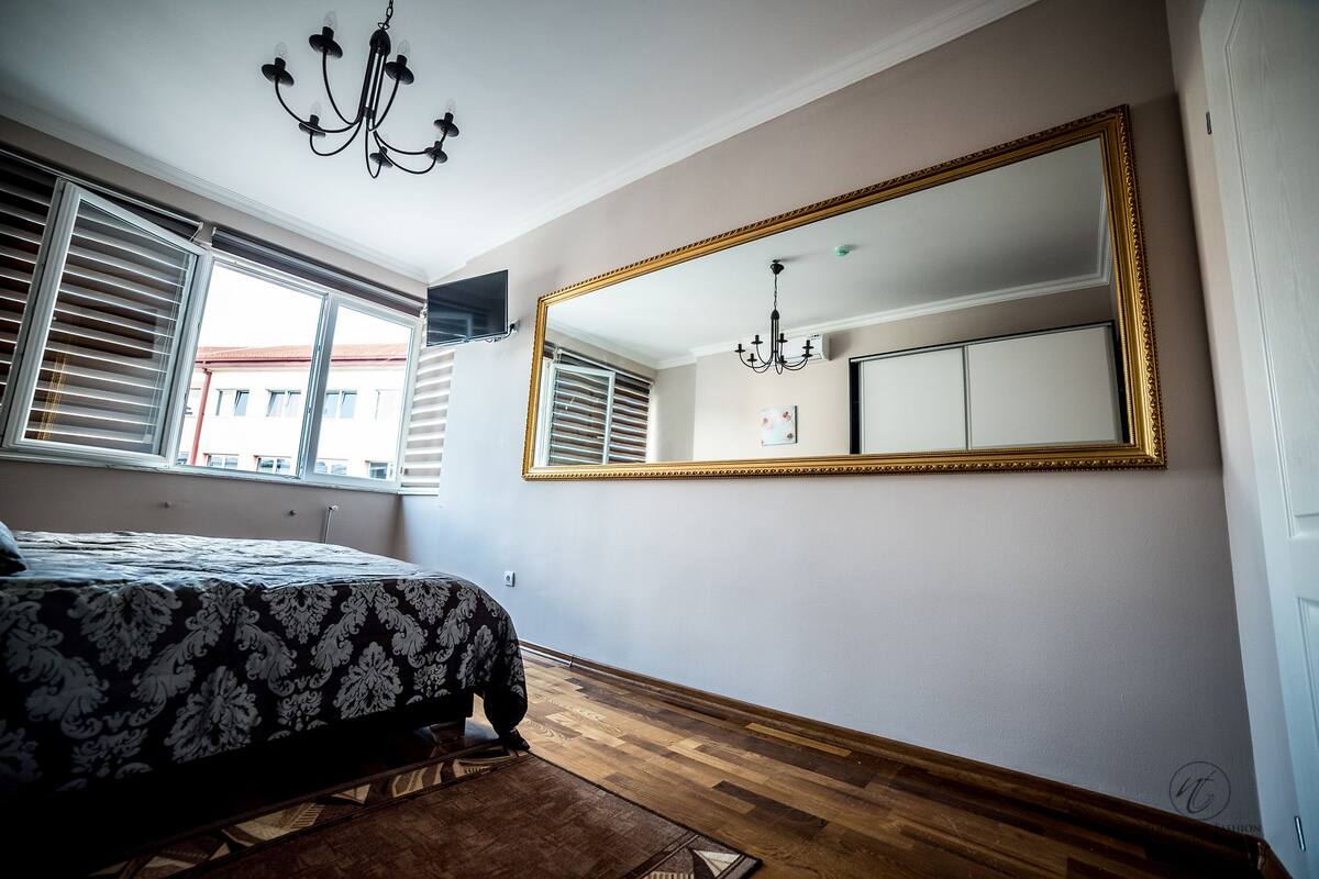 Un dormitorio con un espejo grande, una cama con edredón estampado, suelo de madera y una araña.