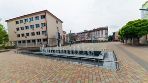 A public square with a fountain, a multi-story building, and a tree. - Suite Donna (Bihac)