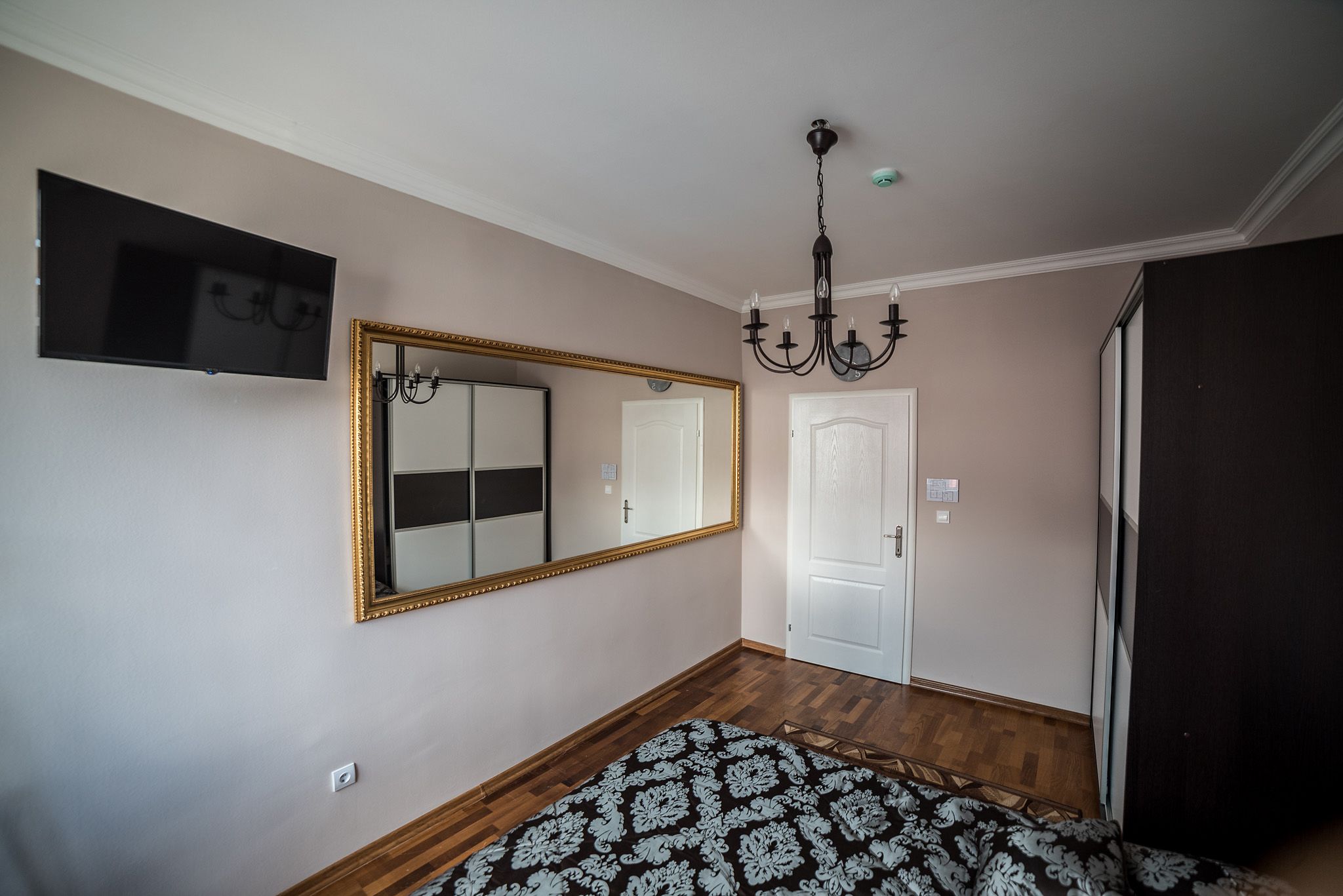 A bedroom with a large mirror, a chandelier, a flat-screen TV, and a patterned rug.