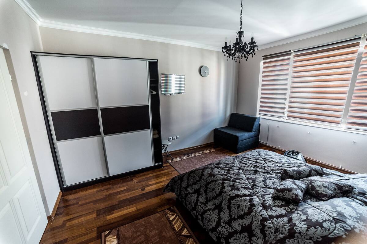 Un dormitorio con cama, armario, silla, ventana con persianas, reloj y lámpara de araña.