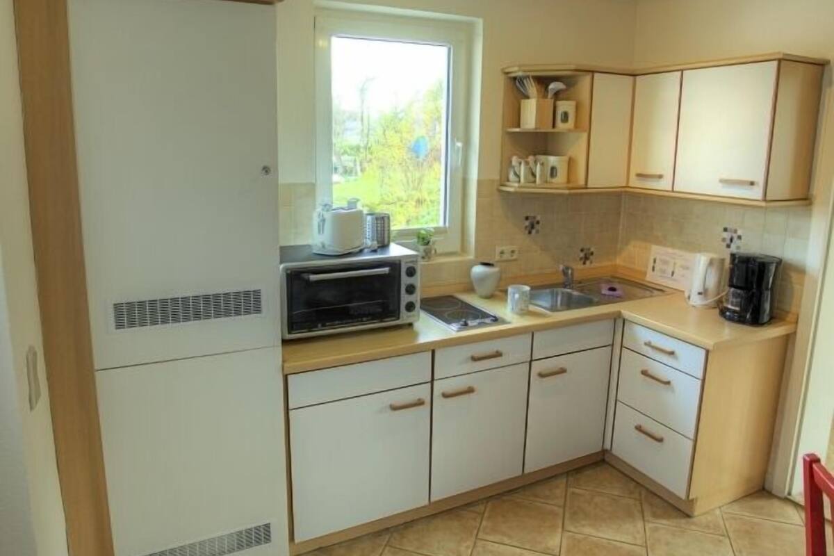 Eine kleine Küche mit Einbauofen, Mikrowelle und Spüle. Ein Fenster sorgt für natürliches Licht, und die Küche hat ein schlichtes, funktionales Design mit hellen Schränken und einem Fliesenboden.