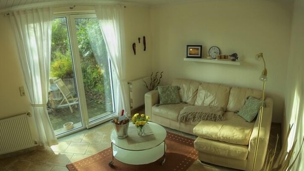 A living room with a beige sofa, a white coffee table, and a brown rug. There is a window with curtains, a radiator, and a plant on the windowsill. - Fewos am Hafen (Sellin)