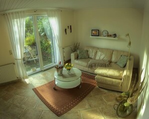 Een woonkamer met een beige bank, een witte salontafel en een bruin vloerkleed. Er is een raam met gordijnen, een radiator en een plant op de vensterbank.