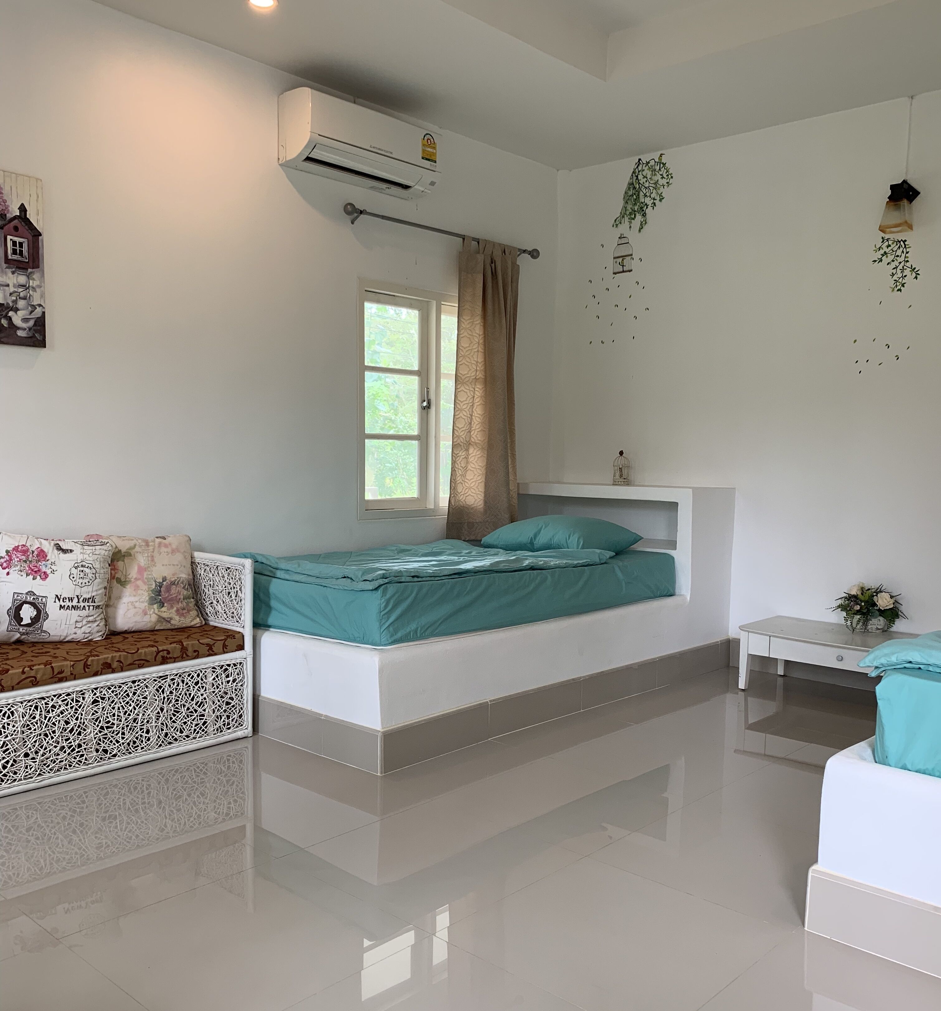 A bedroom with a bed, a window, an air conditioning unit, a small table, and a decorative plant.